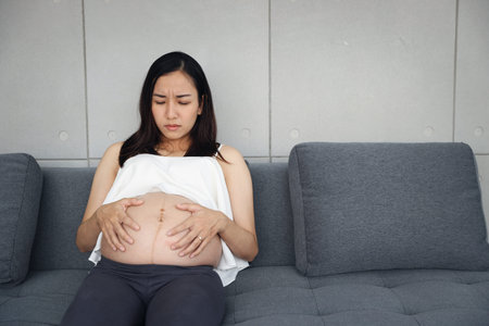 asian pregnant woman is sitting in living room and stressed another hand holding her stomachの写真素材