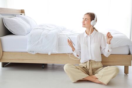 Young attractive caucasian woman wearing white casual clothes with headphone. She listening music or radio by using cell phone or music player in bedroom. (relaxation with technology concept)の写真素材