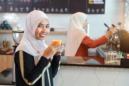 muslim customer drinking coffee at counter with young muslim barista girl blurred backgroundの写真素材