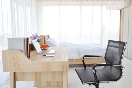 working desk with computer, cell phone and a cup of coffee in white bed room with bedの写真素材