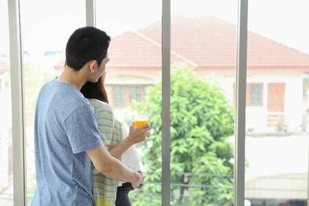 back view image of young asian husband embracing young asian pregnant belly wife and looking at something through the window with happiness and smile while her holding orange juiceの写真素材