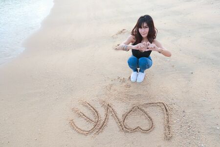 portrait of beautiful woman doing heart symbol on hands with love word on sandの写真素材