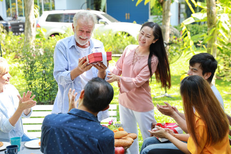 Happy multiethnic family giving surprise gift to caucasian grandfather on his happy birthday with happy smiling face  in backyard outdoor on sunny dayの写真素材