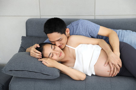 young husband lie down on sofa and embracing and kissing pregnant belly wife with love and happiness...