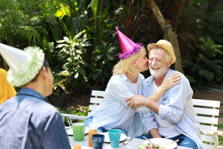 Happy multiethnic family sitting at a breakfast table in backyard outdoor on sunny day with smiling face on birthday or wedding anniversary day. Caucasian elder wife kissing her caucasian husband with laughing.の写真素材
