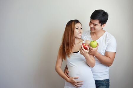 medium shot of young caucasian husband embracing young asian pregnant belly wife with happiness and smile while their holding red and green apples and eyes contract together in bedroomの写真素材