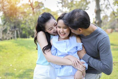 image of happy family, daughter putting her hands on parents cheek with smiling while father and mother kissing during summer time in the parkの写真素材