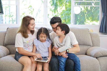 Happy Asian family teaching their childrens son and daughter how to use tablet while sitting on grey sofa in living room with smiling facesの写真素材