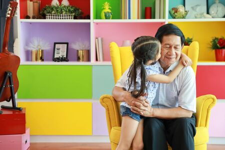 asian granddaughter pecking grandfather on his cheek and pose for a photo with love and happy face on sofa in living roomの写真素材