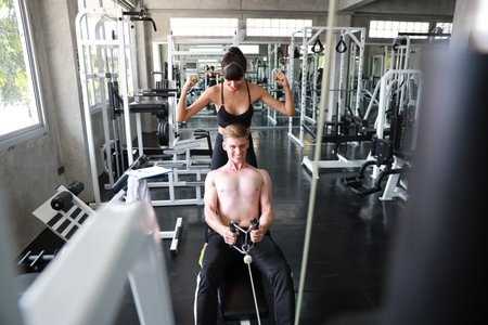 sporty caucasian fitness man using exercise machine in gym with caucasian trainer woman