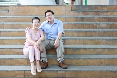 asian traveler couple husband and wife in pink and blue shirt with white nice hat sitting on the stairs in downtown and take a rest with happy smiling face while embracingの写真素材