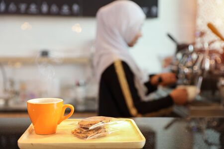 a cup of coffee with cookies at counter with young muslim barista girl blurred backgroundの写真素材