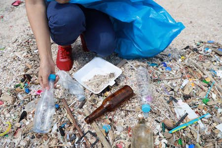 Volunteer tourist with hat is cleaning up garbage and plastic debris on dirty beach by collecting them into big blue bagの写真素材