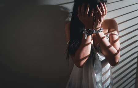 sexual assault and imprisoned asian woman wearing white lingerie sleepwear standing in jail with chain on hands waiting for help (this image for girl victim and violence concept)の写真素材
