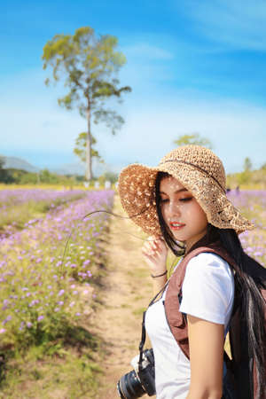 Asian beautiful woman, long hair in white shirt with camera and hat on Verbena filed with blue sky. Portrait cute girl enjoying among flowers farm. Travel in nature outdoor conceptの写真素材