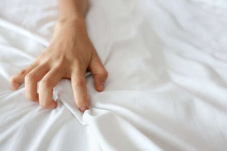 Close up young sexy woman hand pulling white sheets in ecstasy in hotel. Cute girl doing sign orgasm on white bed (sex and erotic concept for advertisement)の写真素材