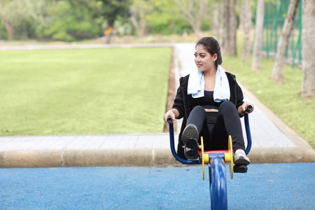 Happy smiling young beautiful asian woman in sportswear relaxing on public exercise machine. Cute...