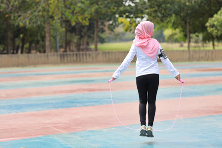Rear view athletic young asian muslim woman in sportswear standing and  skipping rope outdoor for...