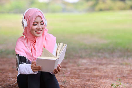 Beautiful fitness athlete asian muslim woman reading book after exercising. Young cute girl sitting in sportswear with mobile phone and headphone taking rest after workout outdoor. Healthy concept.の写真素材