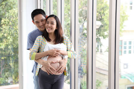 young asian husband standing and embracing young asian pregnant belly wife with happiness and smile in living roomの写真素材