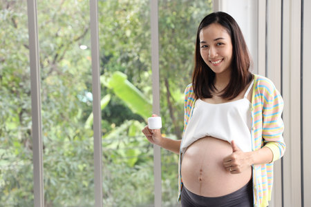 Young asian pregnant belly in casual dress standing and holding cream in living room with happy...