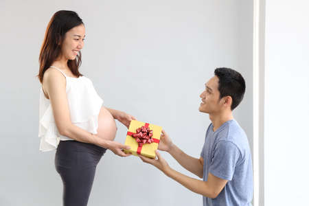 Young asian husband wearing grey t-shirt kneeling and giving presents to young asian pregnant belly wife in living room. Cute girl in white dress smiling while accept gift with happy face.の写真素材