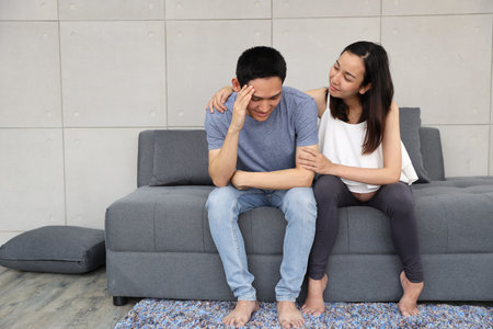 young pregnant wife comforting and touching shoulder young husband while sitting on sofa