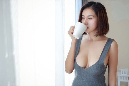 portrait of attractive and sexy asian woman in sexy dress standing beside window and drinking coffee...