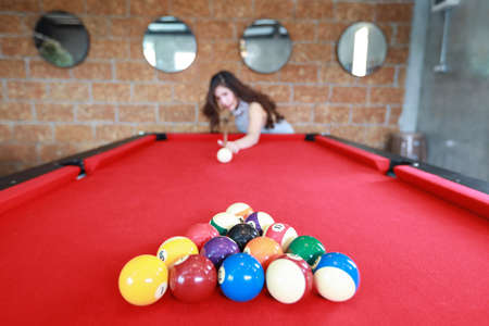 young beautiful asian woman playing and aiming for billiards ball on red pool table in club (focus on balls)の写真素材
