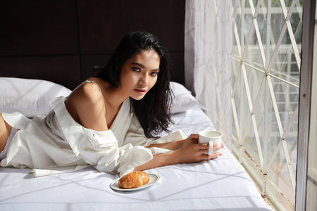 beautiful asian woman in sexy dress having breakfast coffee and croissant while lying in bed and she looking at cameraの写真素材