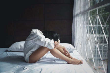 side view asian woman in white lingerie with problem, she bent her head while sitting on bed in bedroom with serious emotionの写真素材