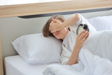 Upset young caucasian woman in white sleepwear lying in bed, wake up late and overslept morning,...