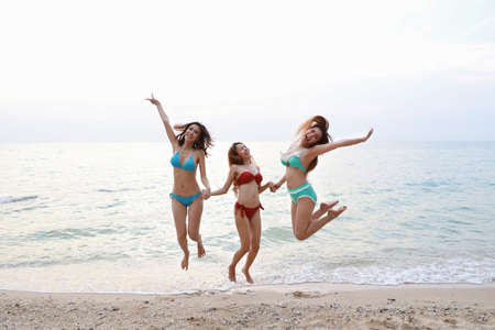 full shot of young asian beautiful and joyful girls in bikini who jumping and raising hands on tropical beach with happy and smile faces on holiday vacation with blue sky (party and friendship concept)の写真素材