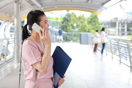 side view successful elder asian businesswoman in pink casual dress who standing on walk way in downtown, she using cell phone and holding folder with city backgroundの写真素材