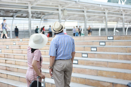 asian traveler couple husband and wife in pink and blue shirt with white nice hat and luggage walking in downtown, he pointing something with left copy spaceの写真素材