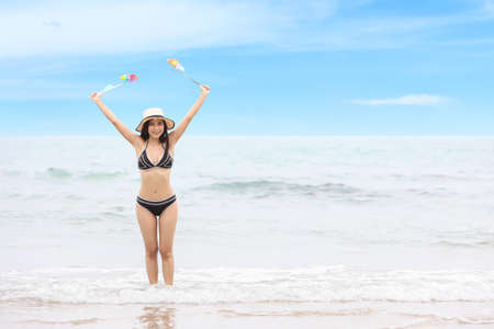 young asian beautiful and sexy girl in black bikini who standing and holding rainbow colored windmills toy and hands up with happy and smile face with blue sky on holiday vacation (party and friendship concept)の写真素材