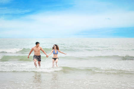 cheerful asian couple wearing blue bikini and black swimsuit. They having fun while holding hands and walking or running on the beach with love on honeymoon day during summer with clear blue skyの写真素材