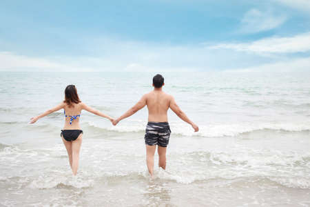 Back view of cheerful asian couple wearing bikini or swimsuit. They having fun while holding hands and walking or running on the beach with love on honeymoon day during summerの写真素材