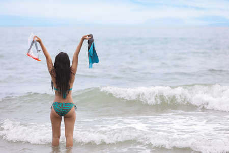 Back view of sexy and beautiful girl who wearing green bikini and holding blue scuba diver and orange snorkel on tropical beach in summer time with right copy spaceの写真素材