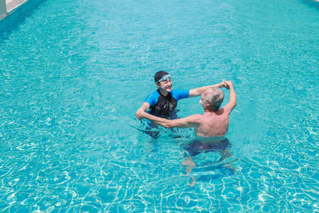 happy elderly caucasian husband and elderly asian wife swimming in pool during retirement holiday with relaxation and happily on sunny day outdoorの写真素材