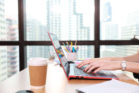 close up and side view of beautiful intern woman hands working or typing on black computer screen with office supplies and coffee cup on wood desk in modern office with city blurred backgroundの写真素材