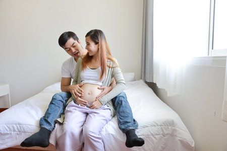 young caucasian husband embracing young pregnant asian belly wife on bed in bedroom with happy...