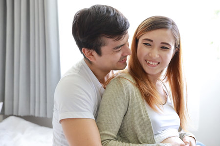 close up young caucasian husband embracing young pregnant asian belly wife on bed in bedroom