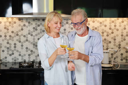 Couple senior caucasian husband and wife in casual dress standing, raising drink for cheers celebration in kitchen at house. Happy elderly man and woman holding glass of wine or cocktails with smilingの写真素材