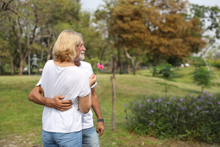side view of elderly caucasian couple standing and embracing while holding red rose in park in...