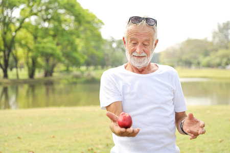 elderly husband wearing sunglasses and white shirt is holding apple in his hand in the park during...