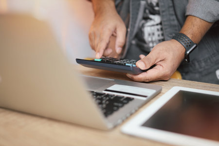 Man hands in grey jean standing and using calculator and laptop computer on wooden table for online...