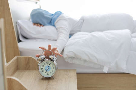 Oversleeping asian muslim woman wearing white sleepwear lying on bed, missing ring of alarm clock waking up. Rear view portrait young girl hand trying to stop ringing while her face down on pillowの写真素材