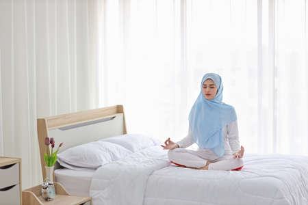 Young asian muslim woman sitting on bed and enjoying meditation. Beautiful girl in sleepwear with blue hijab practices yoga in bedroom with peace and calm. Healthy and lifestyle conceptの写真素材