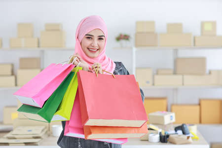Active smart asian muslim woman in jean jacket standing and holding shopping bags with online package box delivery background. Beautiful girl looking at camera and smile. Shopping conceptの写真素材